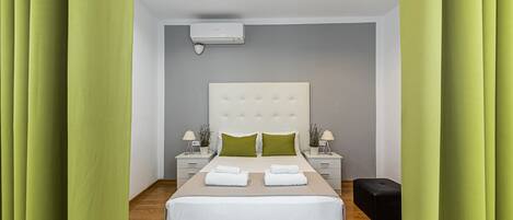 Un dormitorio moderno con una cama grande, almohadas verdes, ropa de cama blanca, dos mesitas de noche con lámparas y un reposapiés negro.