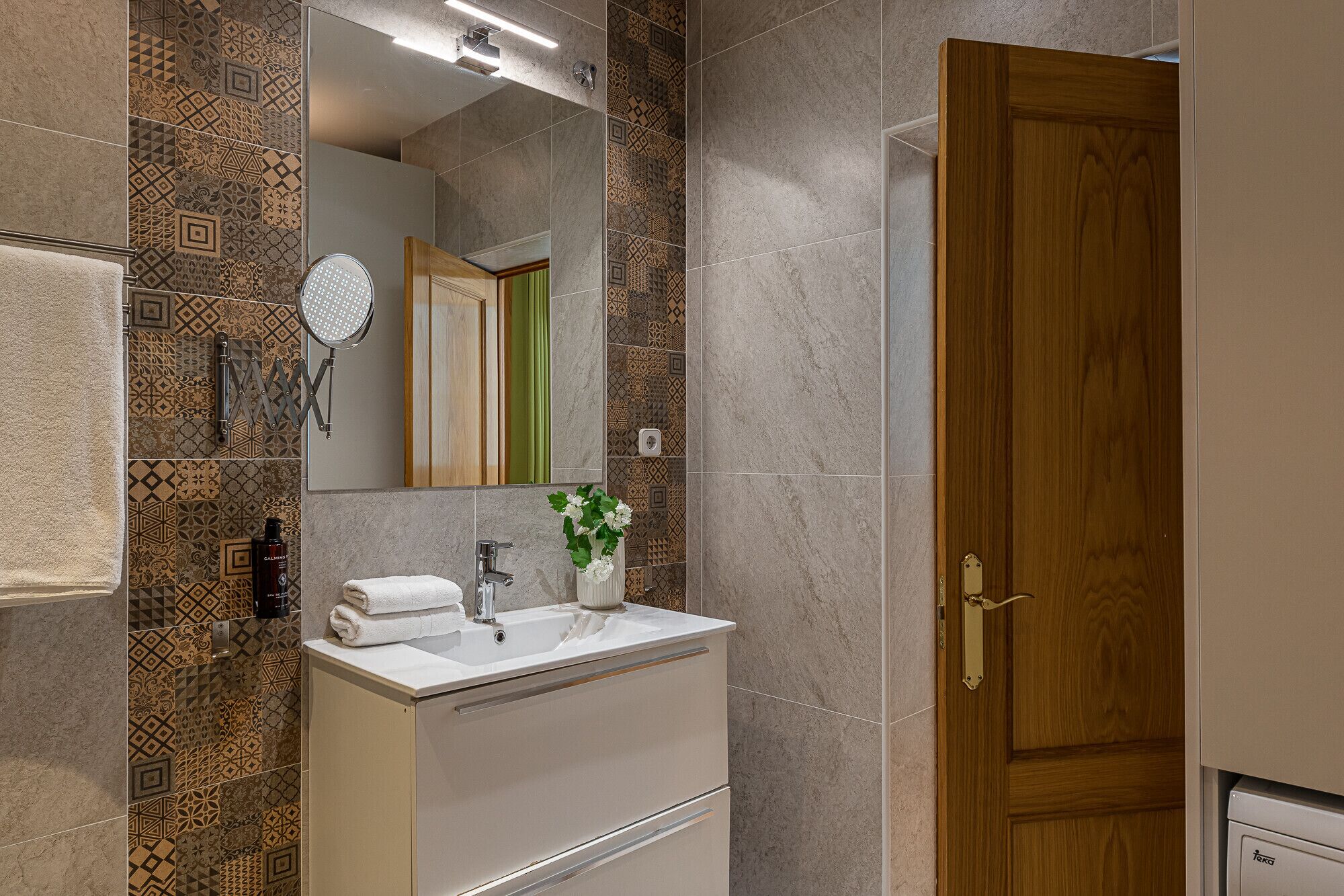 A modern bathroom with patterned tiles, a wooden door, and a white vanity with a mirror and towel rack.