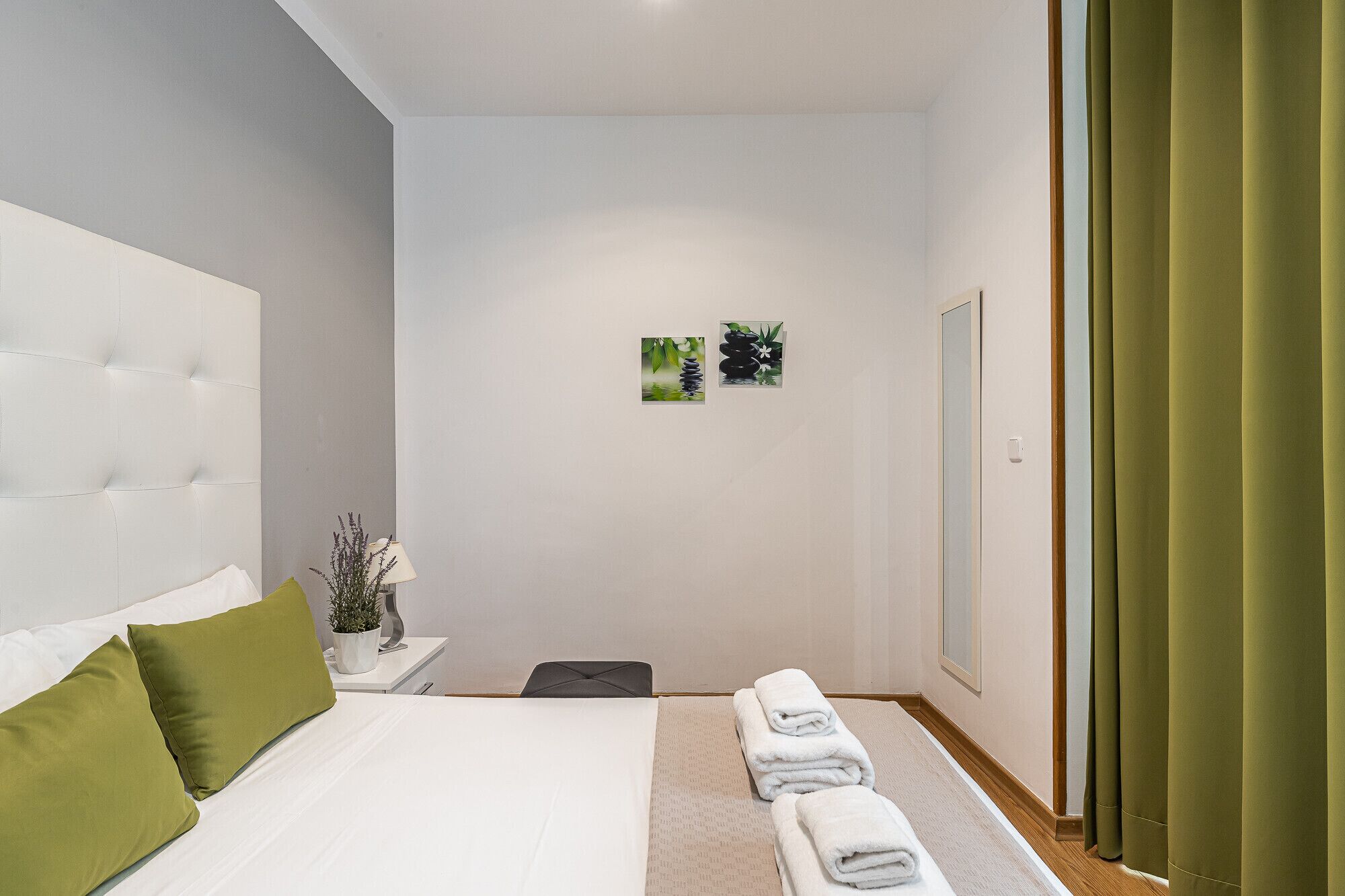 Une chambre d’hôtel avec un grand lit, des oreillers verts, une petite table de chevet avec une lampe et une plante, une œuvre d’art accrochée au mur et un rideau vert.