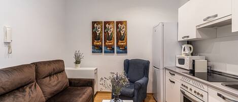 En hyggelig, lille stue med en brun sofa, en blå lænestol, et lille bord med en vase og et tekøkken med mikroovn, brødrister og ovn.