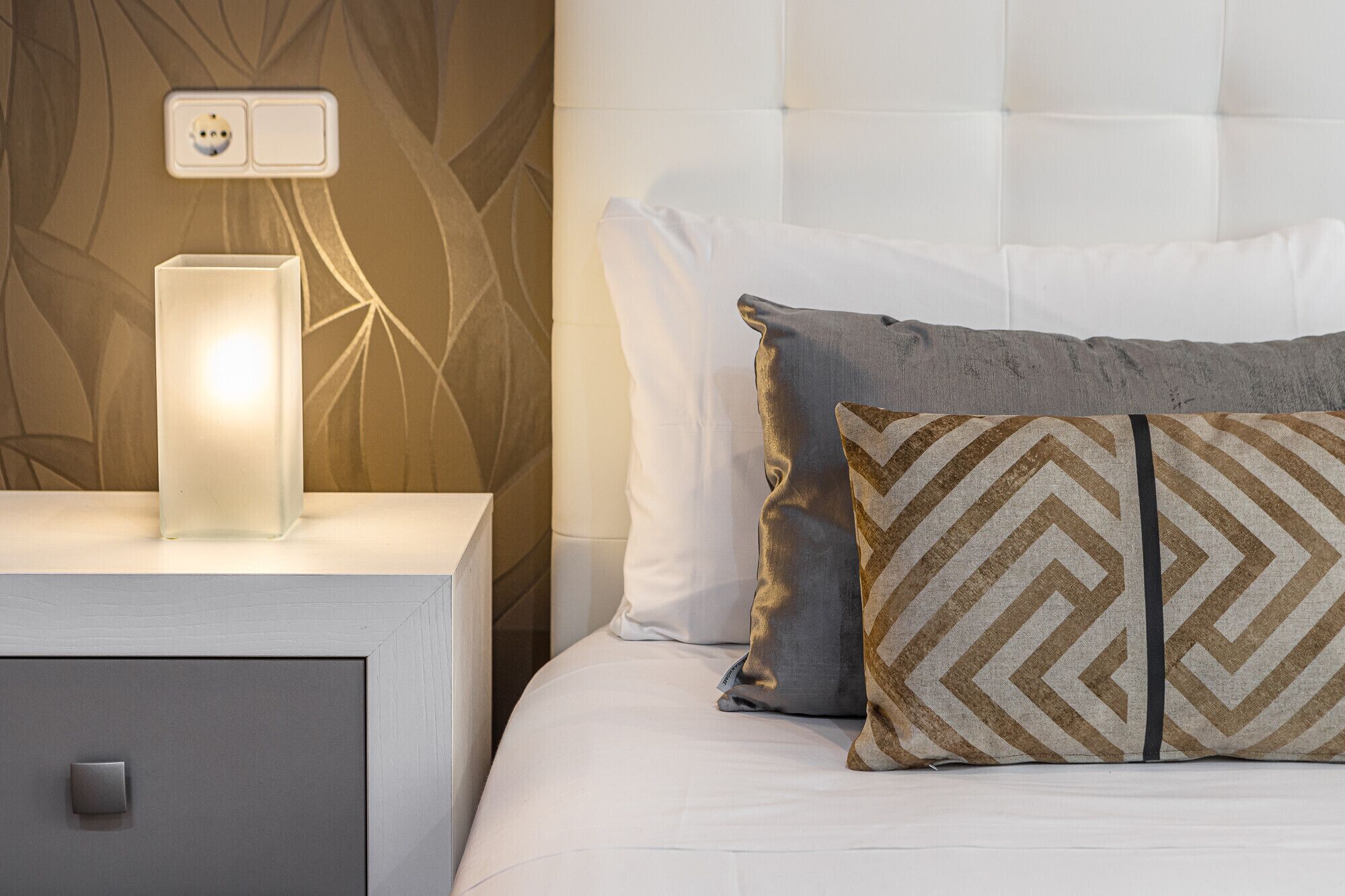 Een opgemaakt bed met een kussen met patroon, een bedlampje en een nachtkastje met een stopcontact.