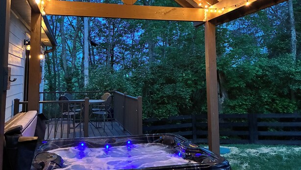 Ein überdachter Außenbereich mit Whirlpool, Holzbalken, Lichterketten und Blick auf Bäume.