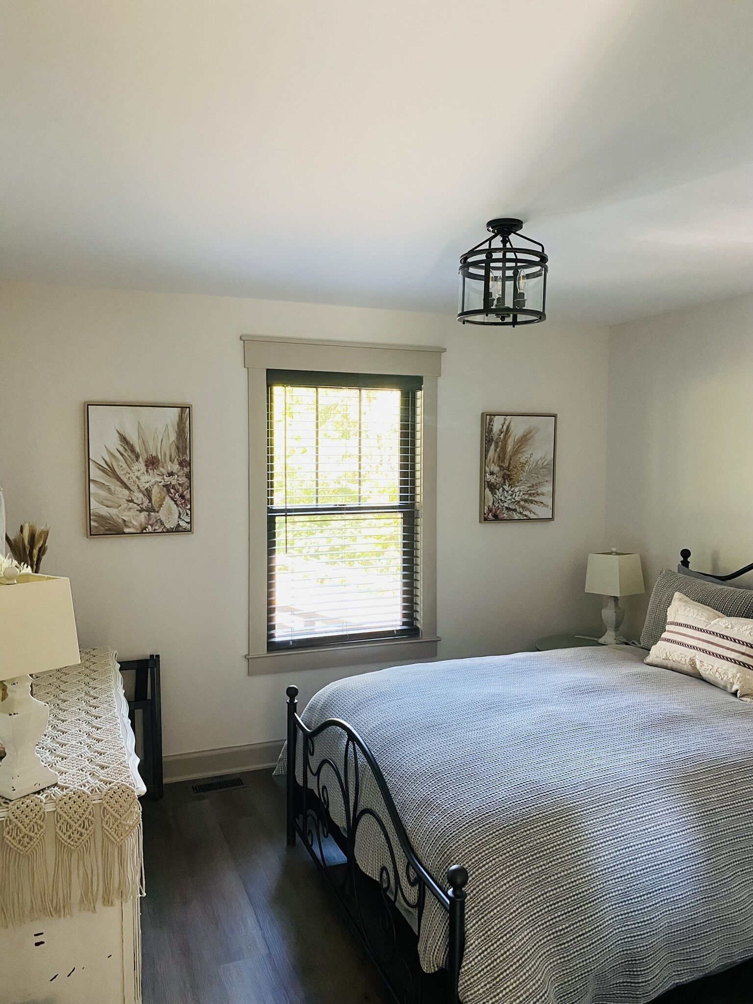 A bedroom with a bed, a window, a nightstand, a lamp, and two wall-mounted paintings.