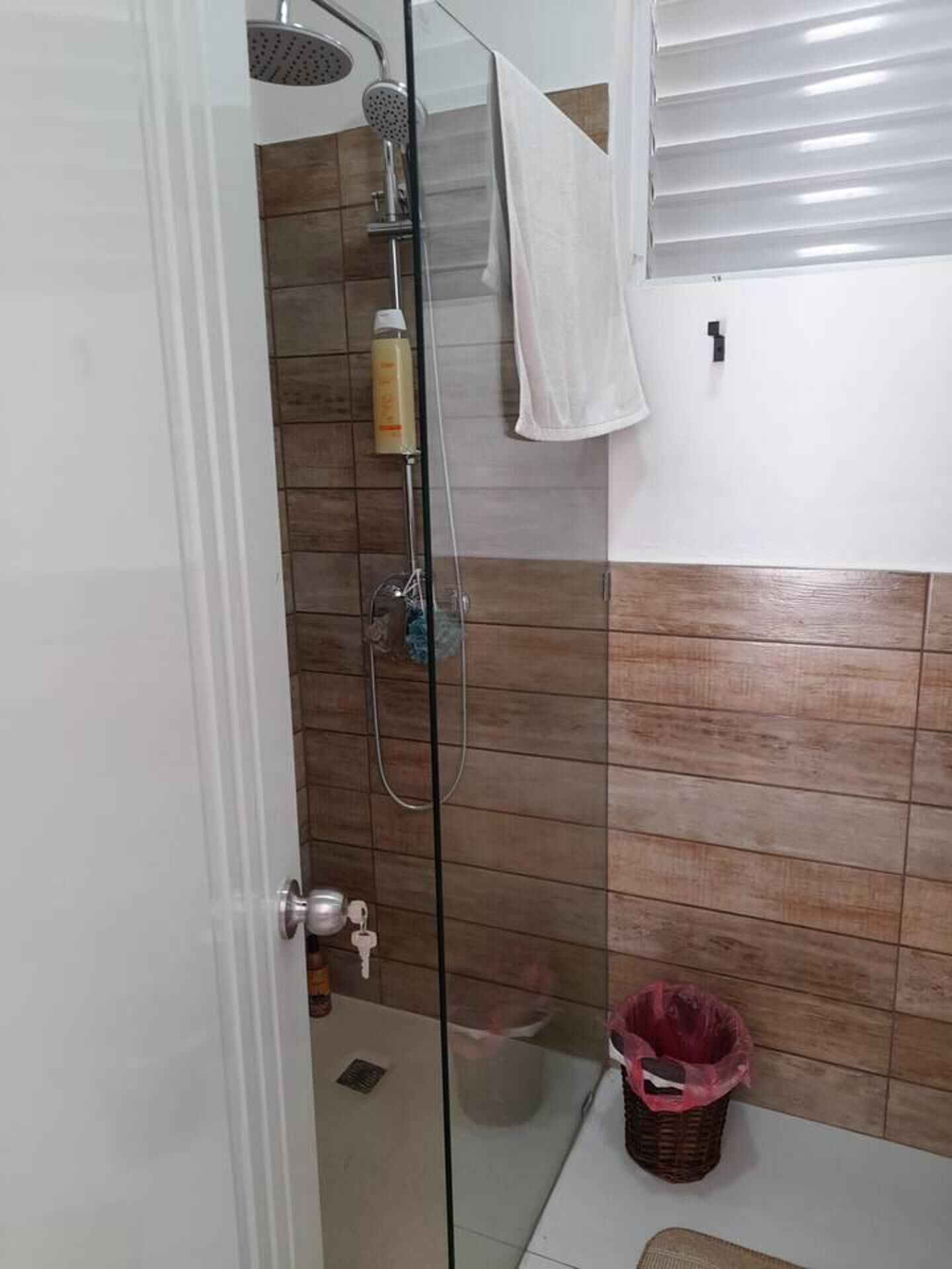 A shower with a glass door, a white towel hanging on the wall, and a trash bin.