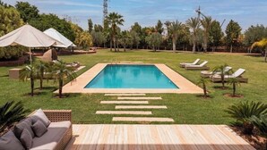 A rectangular swimming pool with a wooden deck in the foreground, surrounded by a well-maintained lawn and palm trees. - Mimouna Riads- Luxury villa 17 bedrooms (Marrakech)