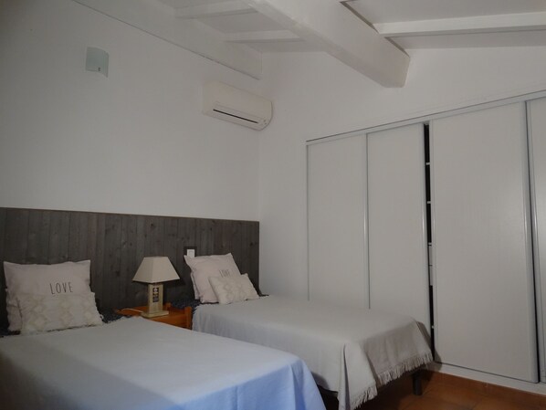 Un dormitorio con dos camas, cabecero de madera, equipo de aire acondicionado, mesita de noche con lámpara y puerta corredera.