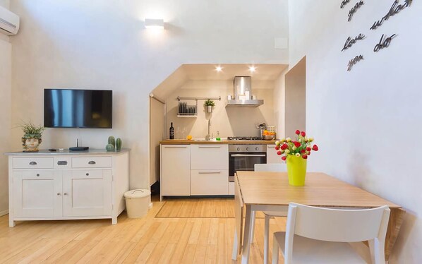 Une cuisine compacte avec un coin repas, des meubles blancs, un téléviseur fixé au-dessus du comptoir et une petite poubelle.