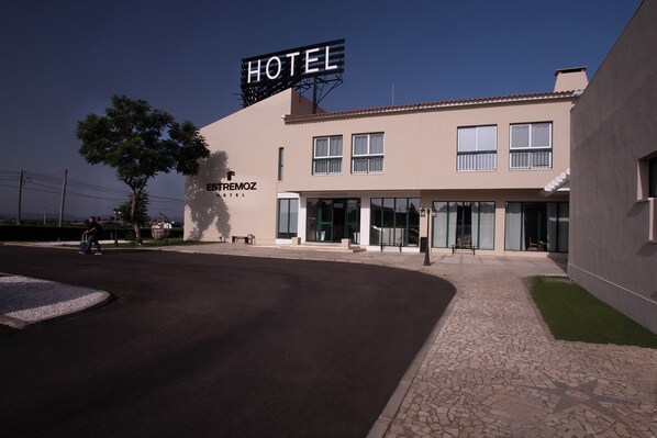 Un hotel moderno con un letrero llamativo, entrada pavimentada y un pequeño espacio al aire libre con mesas y sillas.