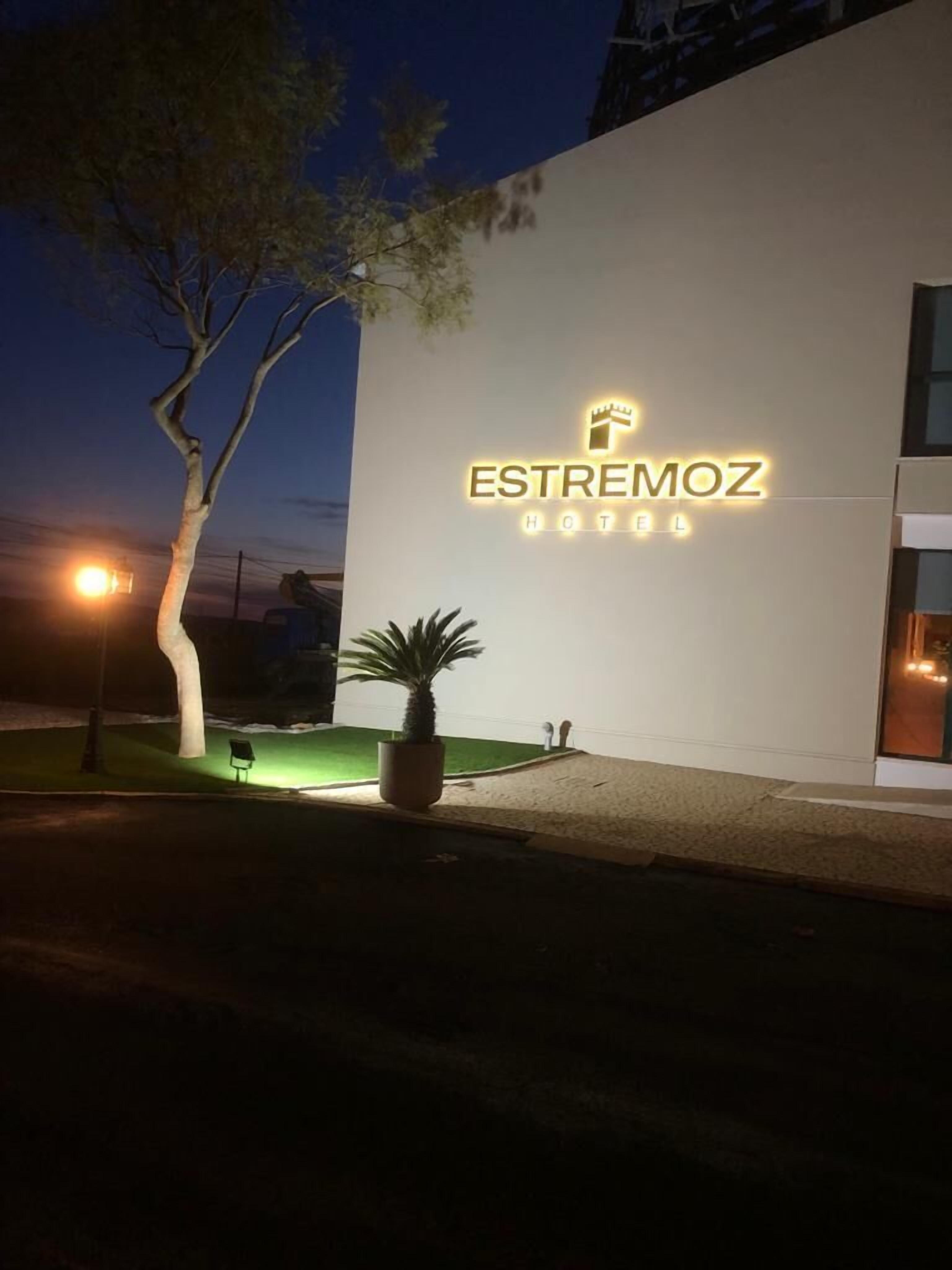 The exterior of Estremoz Hotel with a modern design, a well-lit sign, and a small lawn area with a potted plant.