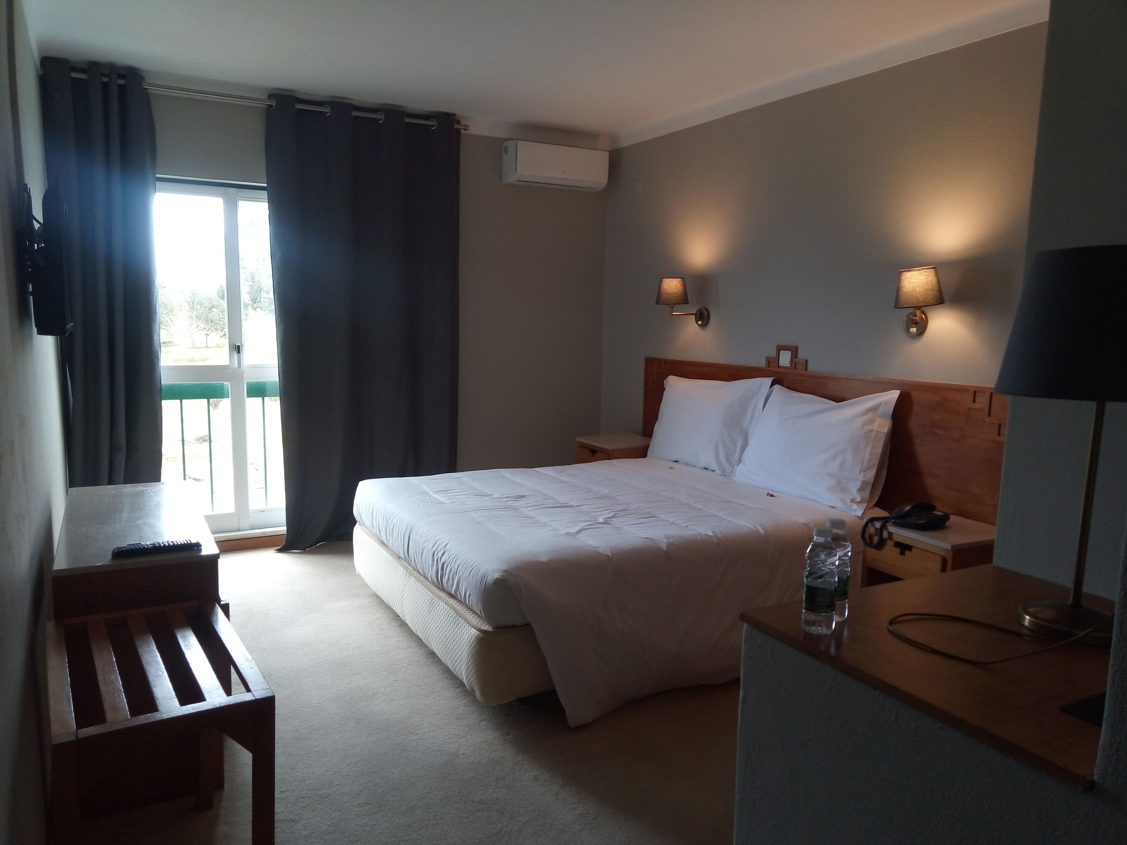A hotel room with a bed, bedside tables, a desk, a chair, a window with curtains, and wall-mounted lamps.