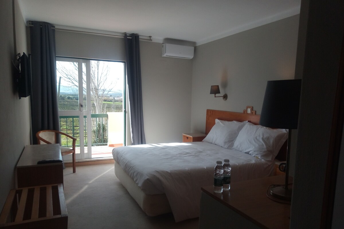 A hotel room with a bed, a desk, a chair, a nightstand, and a view of the outdoors.