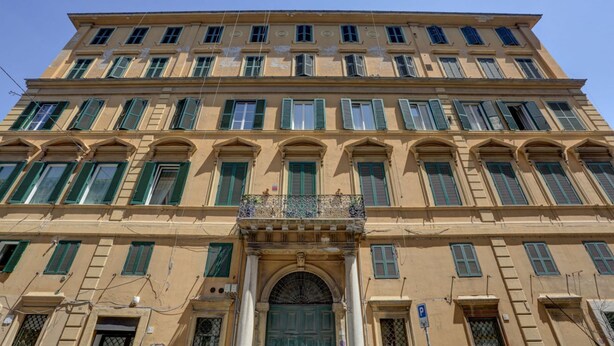 Un immeuble à plusieurs étages doté d’une entrée centrale, de nombreuses fenêtres avec volets et d’un balcon bien visible.