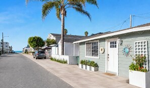 A coastal street with a single-story house featuring a front porch, potted plants, and a decorative clock. - Beach Bungalow Steps to Sand + Hot Tub (Oxnard)