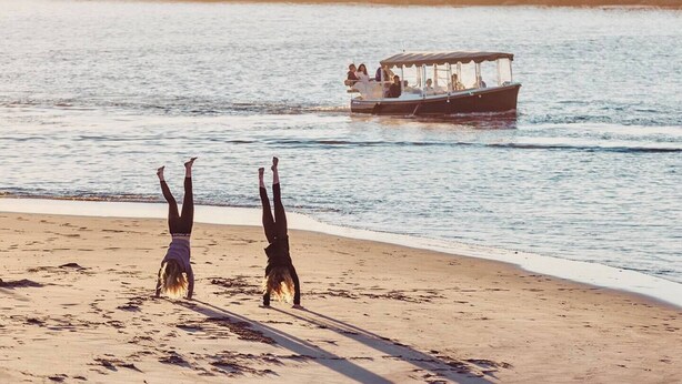 Una spiaggia con due persone che fanno la verticale sulla sabbia, una barca con persone sullo sfondo e una costa rocciosa.