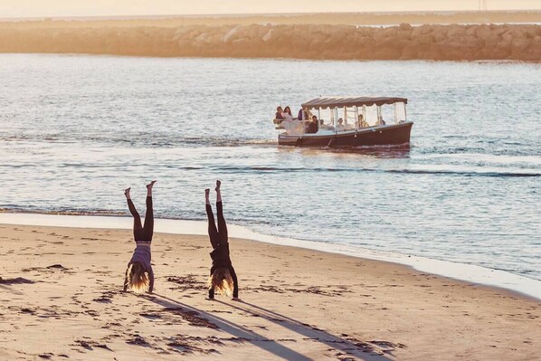 A beach with two people doing handstands on the sand, a boat with people on it in the background, and a rocky shoreline.