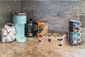 A kitchen counter with a Keurig coffee maker, a coffee pot, a box of coffee pods, a wooden box with coffee cups, and a mug rack.