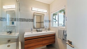Un bagno moderno con cabina doccia in vetro, lavabo doppio con pensili in legno, uno specchio grande e una finestra vista palme.