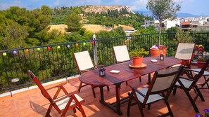 Une terrasse avec une table et des chaises en bois, des plantes en pot et une vue sur une colline, avec des bâtiments visibles au loin.