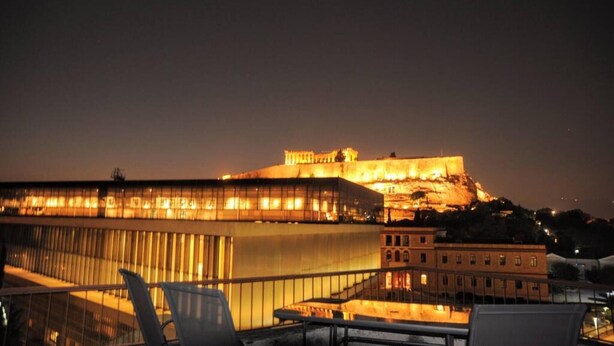 A nighttime view of a historic building with a large, illuminated structure on a hill, overlooking a modern building with a glass facade and outdoor seating area. - Acropolis museum grand apartment (Athina)