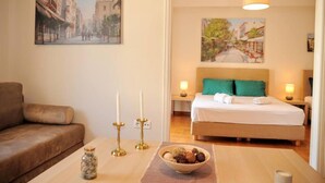 A hotel room with a bed, a nightstand, a table with a bowl of nuts, a candle holder, and a painting of a street scene. - Acropolis museum grand apartment (Athina)