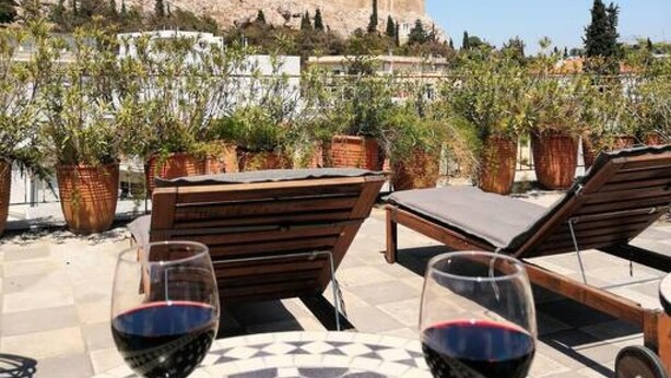 A terrace with two round tables, each with a glass of red wine, wooden lounge chairs, and potted plants. In the background, there is a view of a historical building on a hill. - Stunning Acropolis view apartment (Athina)