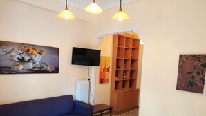 Una stanza con un divano blu, un letto con lenzuola bianche, una TV fissata al muro, un termosifone e un armadio in legno con ripiani.