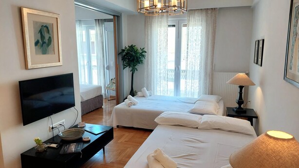 A hotel room with two beds, a TV, a desk with a lamp, and a large window with curtains. - Elegant apartment in Acropolis (Athina)