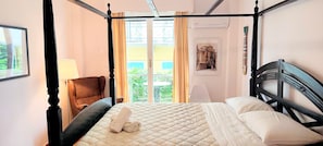 Una cama con dosel con una colcha acolchada, una almohada blanca, una toalla enrollada y una silla con lámpara.