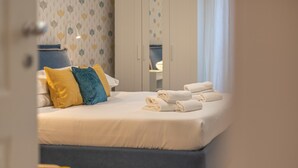 A neatly made bed with white linens and a few pillows, a set of towels on the bed, and a wardrobe in the background.