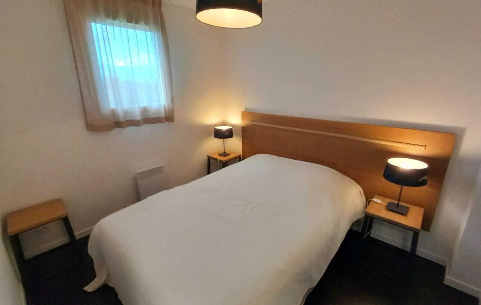 A hotel room with a large bed, two bedside tables with lamps, a window with curtains, and a headboard.