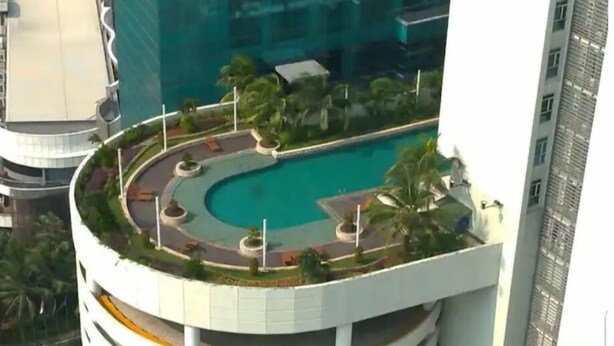 A high-rise building with a rooftop swimming pool surrounded by greenery and palm trees.