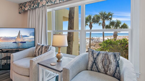 A room with a large window offering a beach view, two armchairs, a small table with a lamp, and a TV displaying a sailboat.