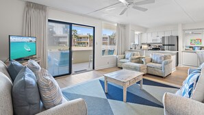 A spacious living room with a large TV, a ceiling fan, and a view of a balcony with buildings in the background. - Freshly renovated! Beachfront property! (Destin)