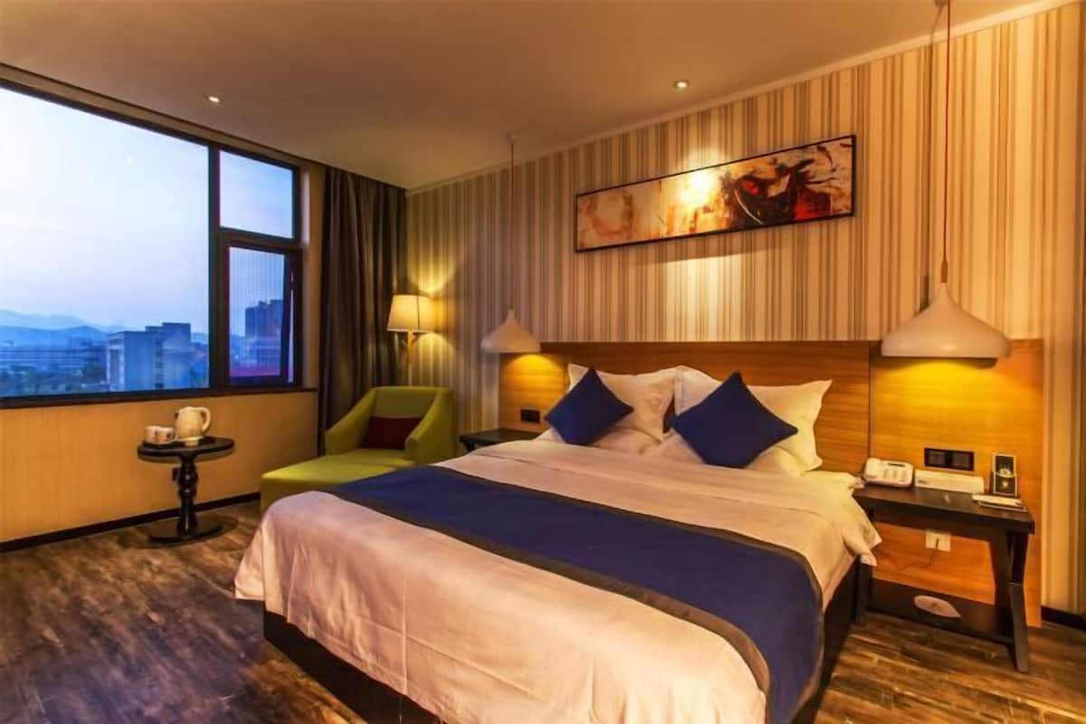 A hotel room with a large bed, a desk, a chair, a window with a city view, and a painting on the wall.