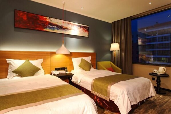 A hotel room with two beds, a large window, a painting on the wall, and a lamp on a side table.