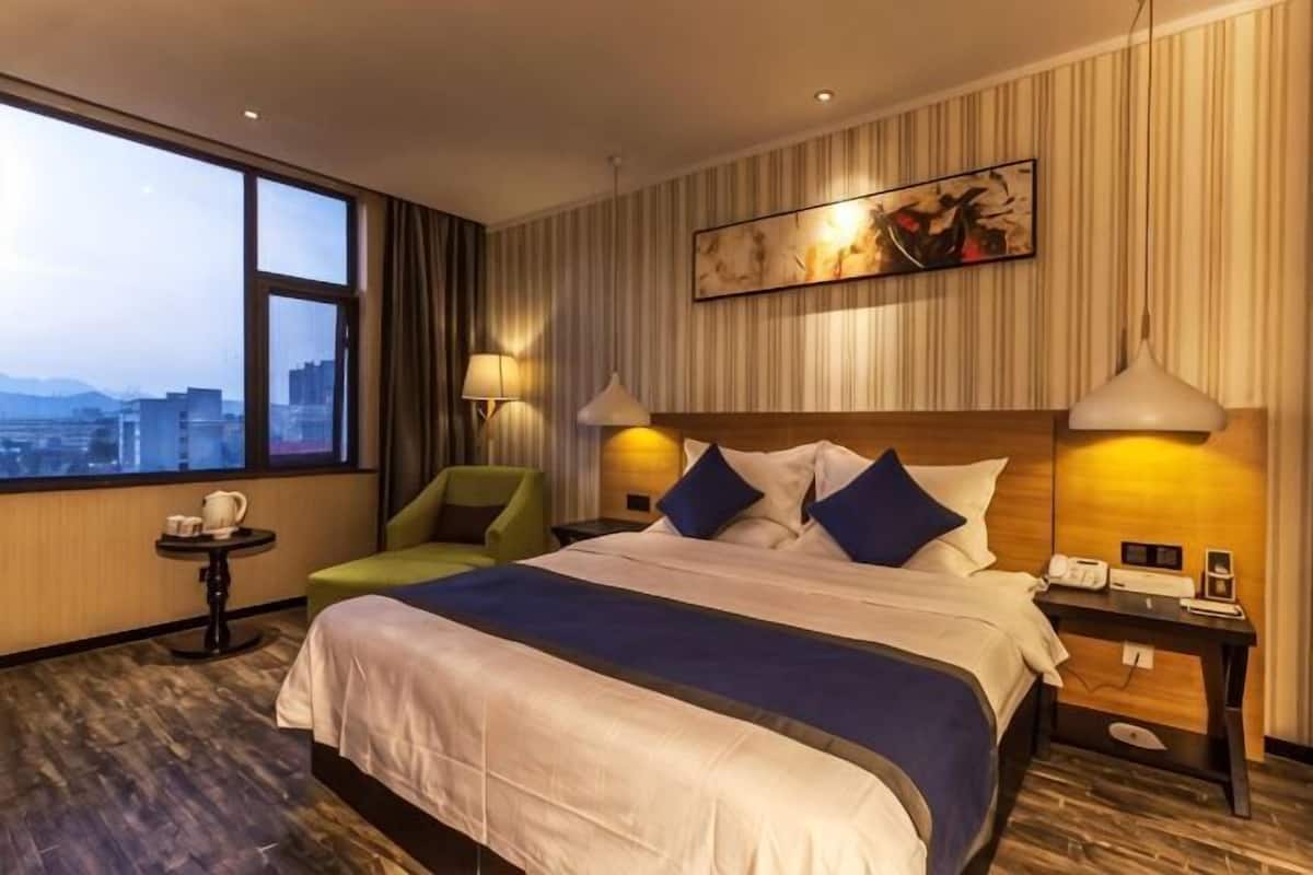 A hotel room with a large bed, a city view, a small table with a lamp, and a chair.