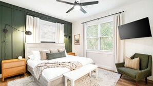 A bedroom with a bed, bedside tables, a bench, a chair, a ceiling fan, a TV, and large windows with curtains. - Top Shenandoah Stay | 4BR w/ 2 King Suites | Private | Easy Drive | Newly Built (McGaheysville)