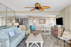 A spacious living room with a light blue sofa, a wooden coffee table, a ceiling fan, and a television on a cabinet.