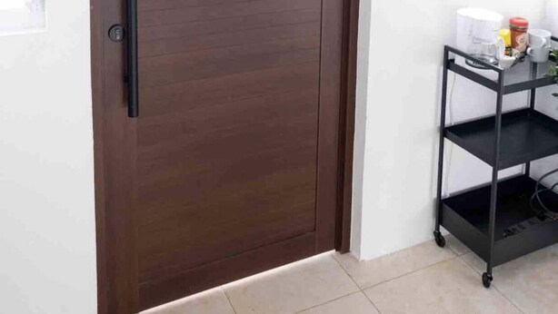 Eine braune, geschlossene Tür mit Griff, ein schwarzes Metallregal mit Gegenständen und ein Fenster mit Rollläden.