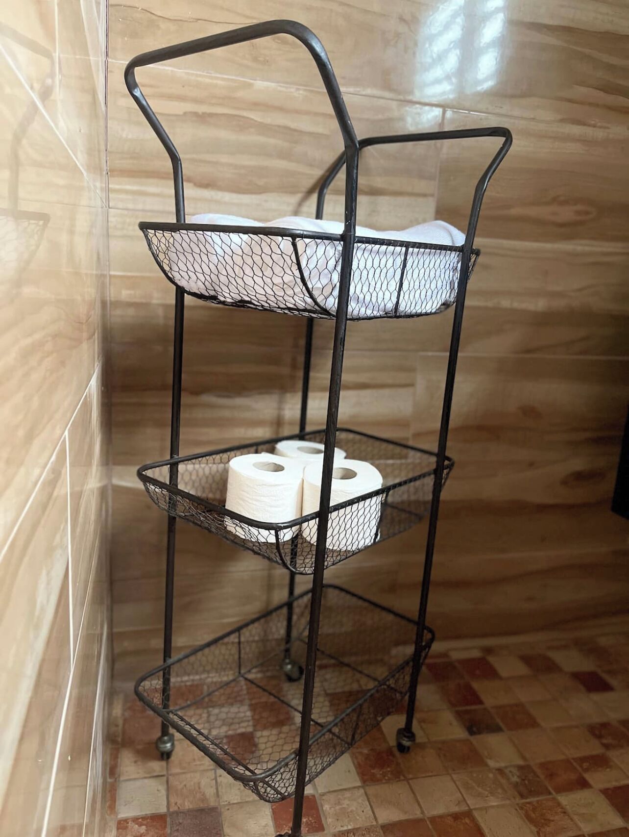 A metal three-tiered shelf with mesh sides, holding toilet paper and a white cloth.