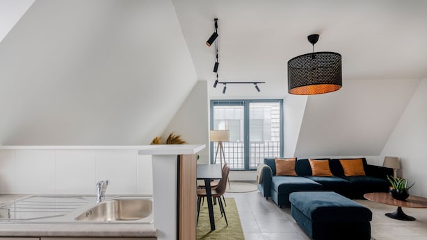 A modern kitchen with a sink and dining area, a dark blue sofa with orange cushions, and a hanging pendant light.