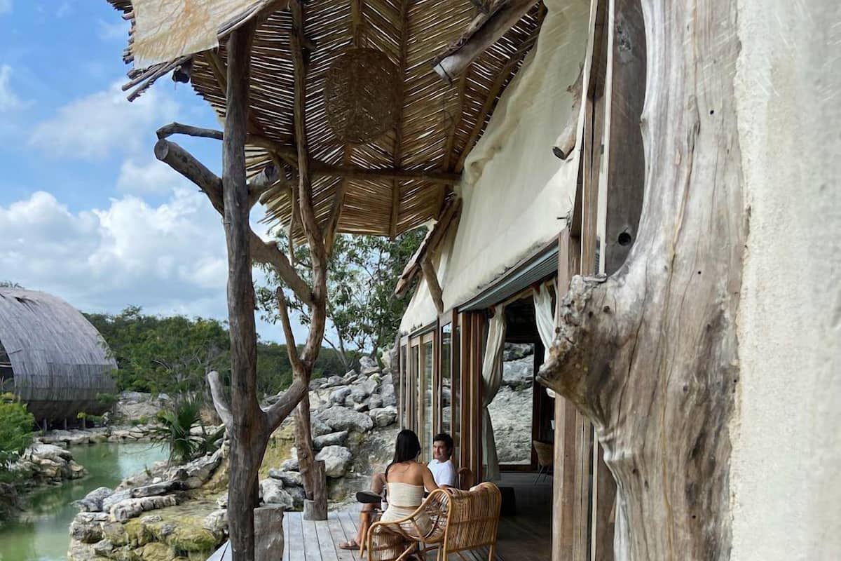 A wooden deck extending over a body of water, with a rustic building featuring large windows and a thatched roof.