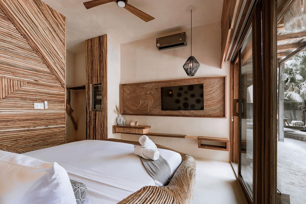 A modern bedroom with a large bed, wooden wall panels, a mounted TV, and a view of the outdoors.