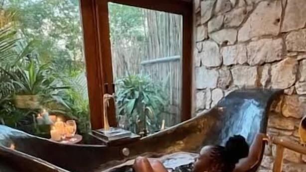 Una habitación con paredes de piedra, un ventanal grande, una bañera de piedra y una silla de madera.