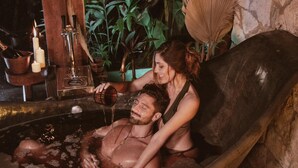 Una pareja en un jacuzzi ovalado y espacioso con decoración rústica, rodeado de plantas y velas.