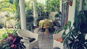 Een overdekt terras met rieten stoelen, een tafel met een groene tafelkleed en potplanten.