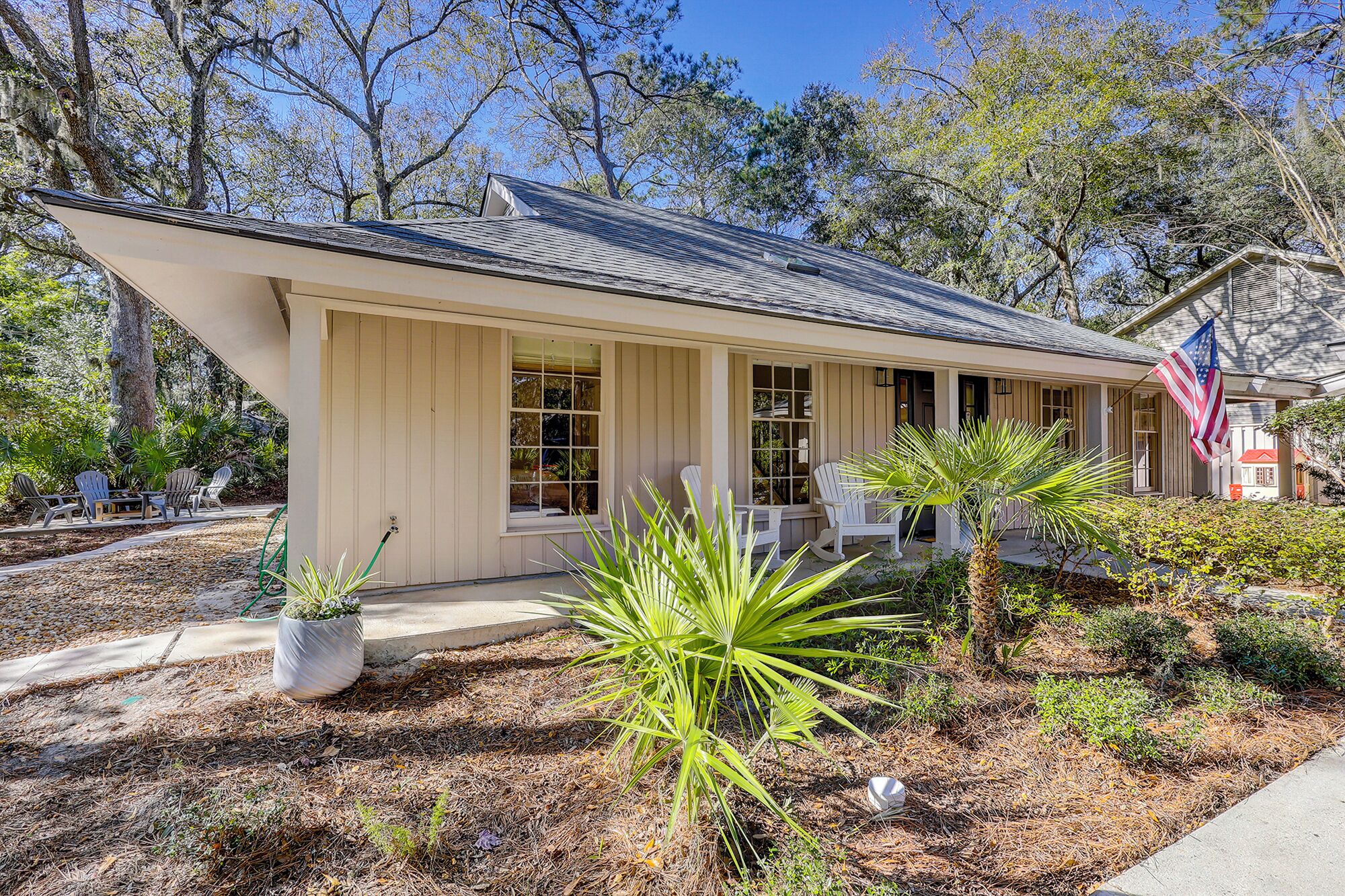 A single-story house with a sloped roof, a porch with white chairs, and a small garden with plants and a palm tree.