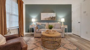 Ein Wohnzimmer mit einem beigefarbenen Sofa, einem runden Korbtisch und einem gemusterten Teppich. Auf weißen Nachttischen stehen jeweils zwei Nachttischlampen.