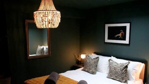 Een slaapkamer met een bed, kussens, een nachtkastje, een lamp, een spiegel en een lijst met een afbeelding aan de muur.