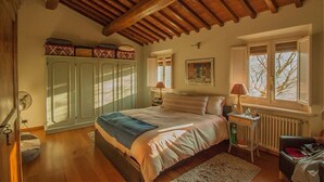 A bedroom with a wooden ceiling, a bed with a blue blanket, a nightstand with a lamp, a radiator, and a window with a view of trees.